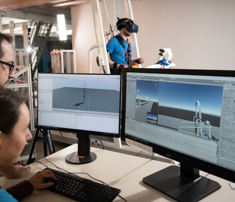 Zu sehen sind zwei Personen vor zwei Bildschirmen, die sich eine Gang-Computersimulation anschauen. Im Hintergrund sieht man die eine Testperson in einem Ganzkörpermessanzug auf einem Laufband. Zu sehen sind zwei Personen vor zwei Bildschirmen, die sich eine Gang-Computersimulation anschauen. Im Hintergrund sieht man die eine Testperson in einem Ganzkörpermessanzug auf einem Laufband.
