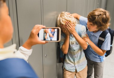 Ein Kind bedrängt ein anderes, welches sich die Hände vor das Gesicht hält. Ein drittes Kind filmt die Situation mit dem Smartphone. Ein Kind bedrängt ein anderes, welches sich die Hände vor das Gesicht hält. Ein drittes Kind filmt die Situation mit dem Smartphone.