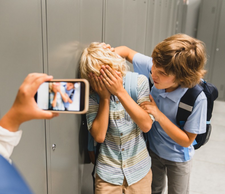 Ein Kind bedrängt ein anderes, welches sich die Hände vor das Gesicht hält. Ein drittes Kind filmt die Situation mit dem Smartphone. Ein Kind bedrängt ein anderes, welches sich die Hände vor das Gesicht hält. Ein drittes Kind filmt die Situation mit dem Smartphone.