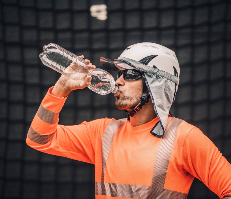 Ein Arbeitnehmer trägt Kleidung zum Schutz gegen Hitze und trinkt Wasser aus einer Flasche. Ein Arbeitnehmer trägt Kleidung zum Schutz gegen Hitze und trinkt Wasser aus einer Flasche.