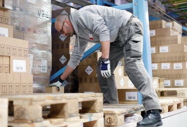 Person in Arbeitskleidung beugt sich in einem Lagerhaus über eine Holzpalette. Im Hintergrund sind Regale mit Kartons zu sehen. Person in Arbeitskleidung beugt sich in einem Lagerhaus über eine Holzpalette. Im Hintergrund sind Regale mit Kartons zu sehen.