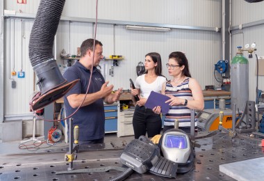 In einer Werkstatt mit metallverarbeitender Ausstattung stehen ein Mann und zwei Frauen im Gespräch. Der Mann trägt ein dunkelblaues Poloshirt und erklärt gestikulierend etwas. Die eine Frau hält ein Klemmbrett, die andere ein Werkzeug. Im Vordergrund liegt ein Schweißhelm auf einem großen Metallarbeitstisch, an dem Absaugvorrichtungen und weitere Werkzeuge angebracht sind. Im Hintergrund sind Werkbänke, Gasflaschen und weitere Arbeitsmittel sichtbar. In einer Werkstatt mit metallverarbeitender Ausstattung stehen ein Mann und zwei Frauen im Gespräch. Der Mann trägt ein dunkelblaues Poloshirt und erklärt gestikulierend etwas. Die eine Frau hält ein Klemmbrett, die andere ein Werkzeug. Im Vordergrund liegt ein Schweißhelm auf einem großen Metallarbeitstisch, an dem Absaugvorrichtungen und weitere Werkzeuge angebracht sind. Im Hintergrund sind Werkbänke, Gasflaschen und weitere Arbeitsmittel sichtbar.