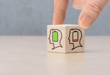 Eine Hand dreht einen kleinen Holzwürfel auf einem Tisch. Auf den sichtbaren Seiten des Würfels sind stilisierte Kopf-Symbole mit Batteriestandsanzeigen abgebildet: einmal mit grün gefüllter Batterie und einmal mit fast leerer roter Batterie. Der Hintergrund ist unscharf und neutral gehalten. (Bild: Celt Studio / stock.adobe.com)
