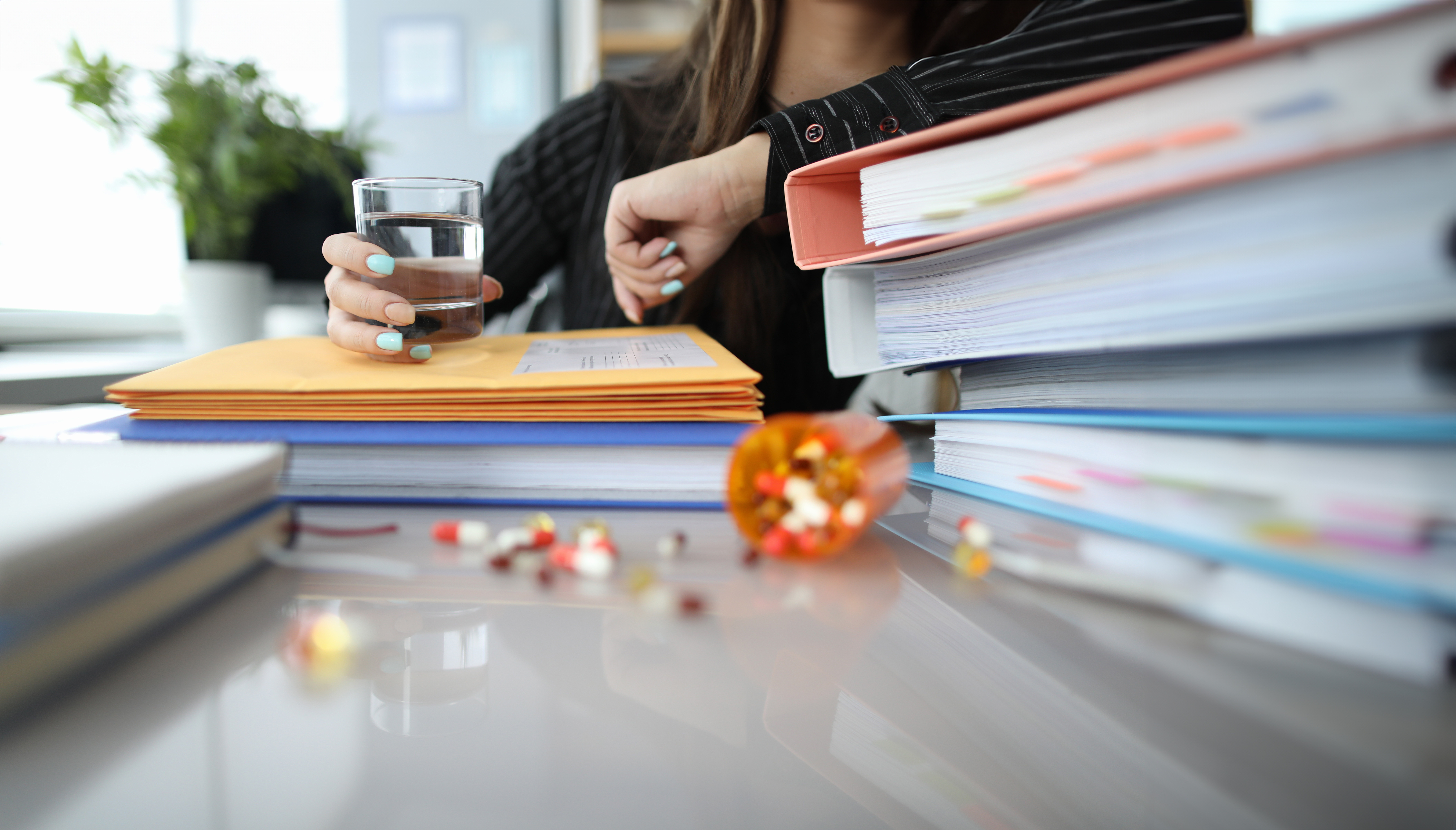 Auf einem Schreibtisch liegen mehrere hohe Stapel von Aktenordnern, Mappen und Dokumenten. Dazwischen sind verstreute bunte Tabletten zu sehen, einige befinden sich noch in einer umgestürzten Medikamentenkapsel. Im Hintergrund hält eine Person ein Glas Wasser in der Hand und stützt sich mit dem Arm auf die Unterlagen. (Bild: H Ko - stock.adobe.com)