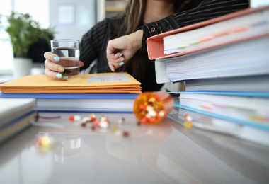 Auf einem Schreibtisch liegen mehrere hohe Stapel von Aktenordnern, Mappen und Dokumenten. Dazwischen sind verstreute bunte Tabletten zu sehen, einige befinden sich noch in einer umgestürzten Medikamentenkapsel. Im Hintergrund hält eine Person ein Glas Wasser in der Hand und stützt sich mit dem Arm auf die Unterlagen. (Bild: H Ko - stock.adobe.com)